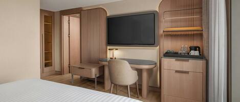 A hotel room with a bed, a desk, a chair, a TV, a minibar, and a wardrobe.