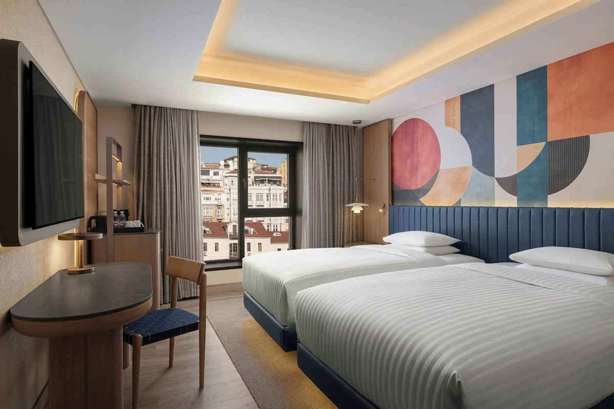 A hotel room with a bed, a desk, a chair, a TV, and a view of buildings.