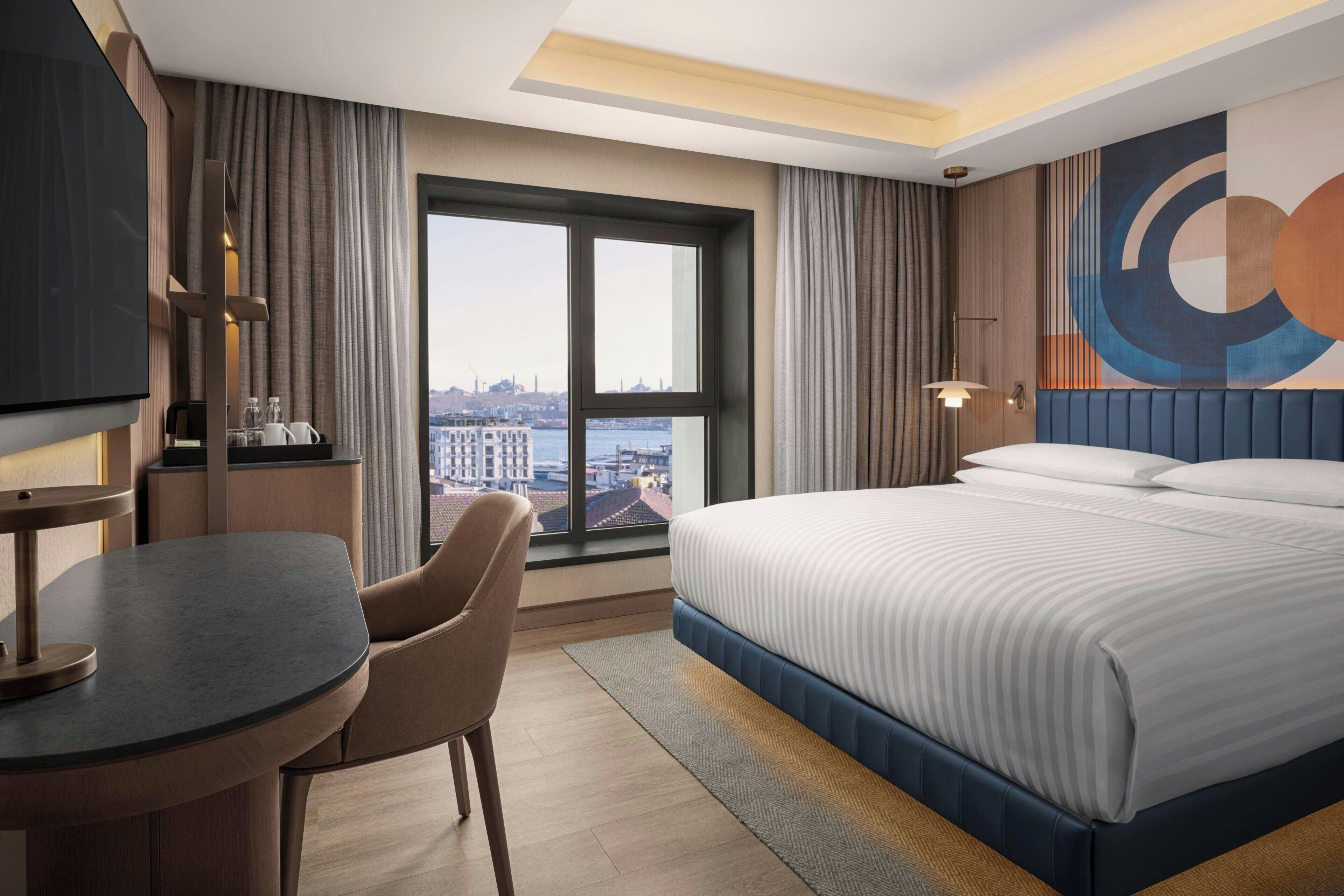 A hotel room with a bed, a desk, a chair, a TV, and a large window with a city view.