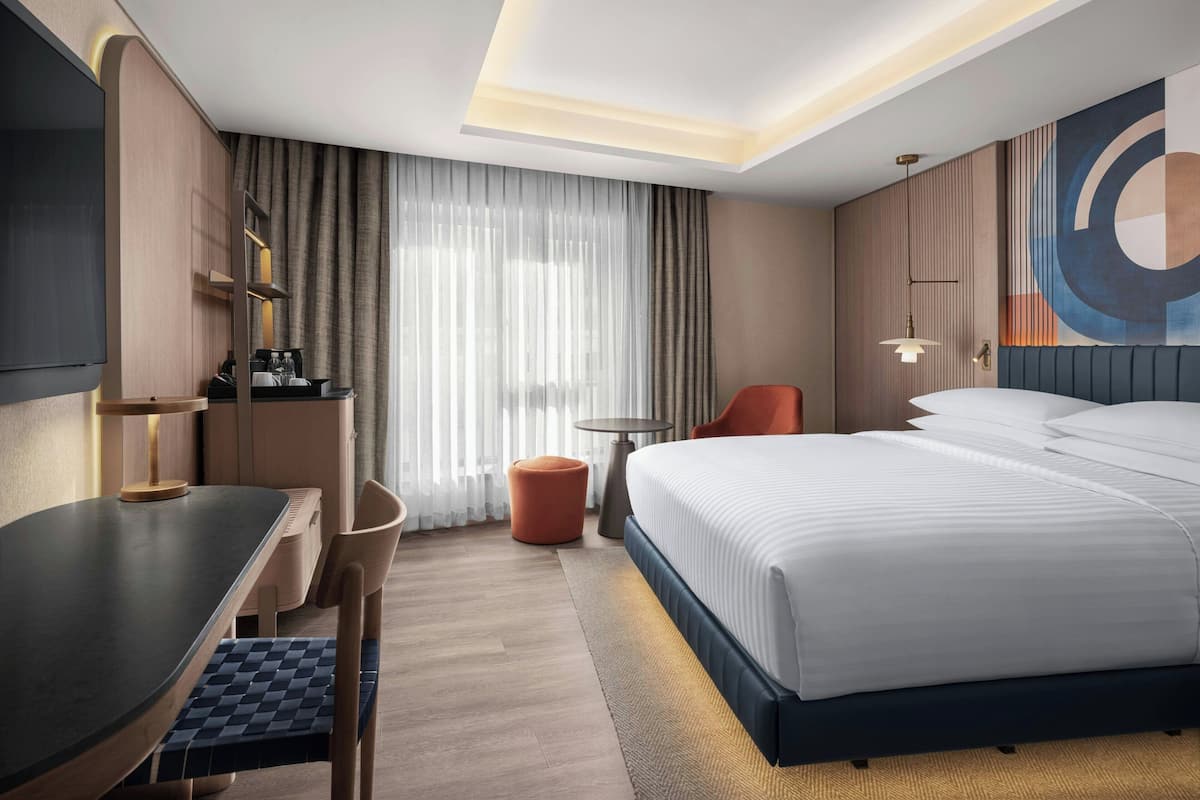 A modern hotel room with a large bed, a desk, a chair, a small table, and a TV.