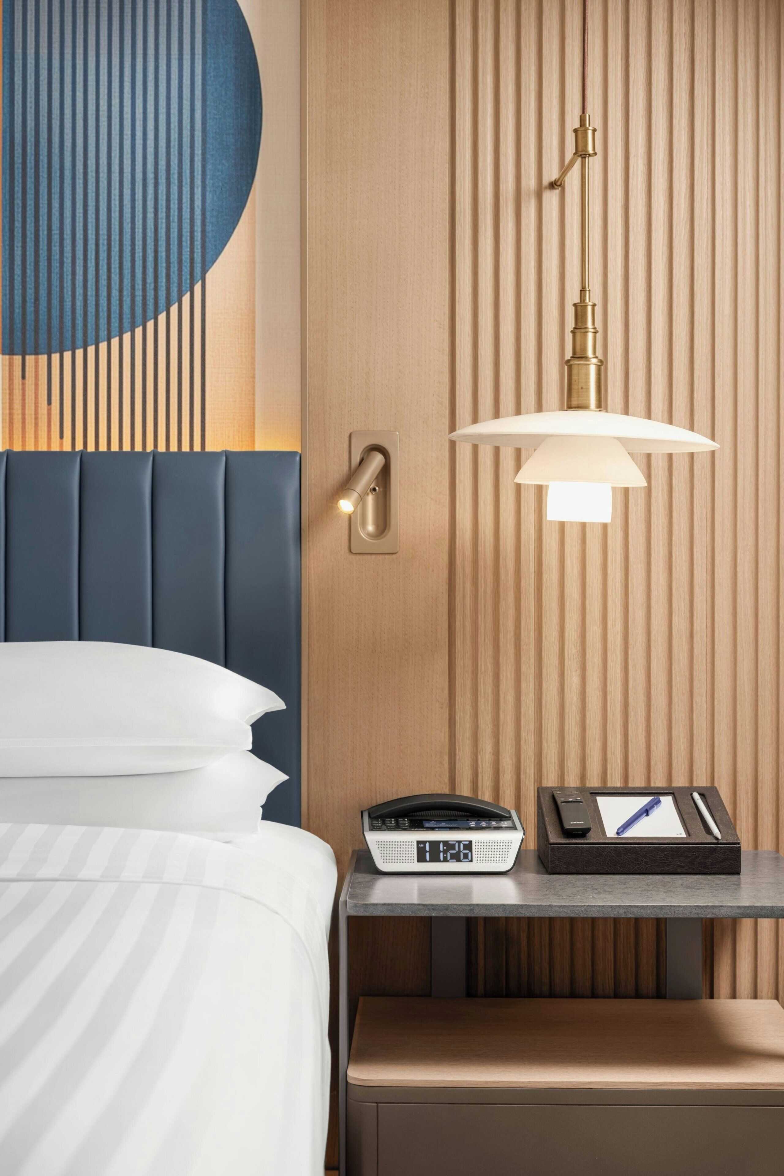 A hotel room with a bed, bedside table, lamp, and a clock.