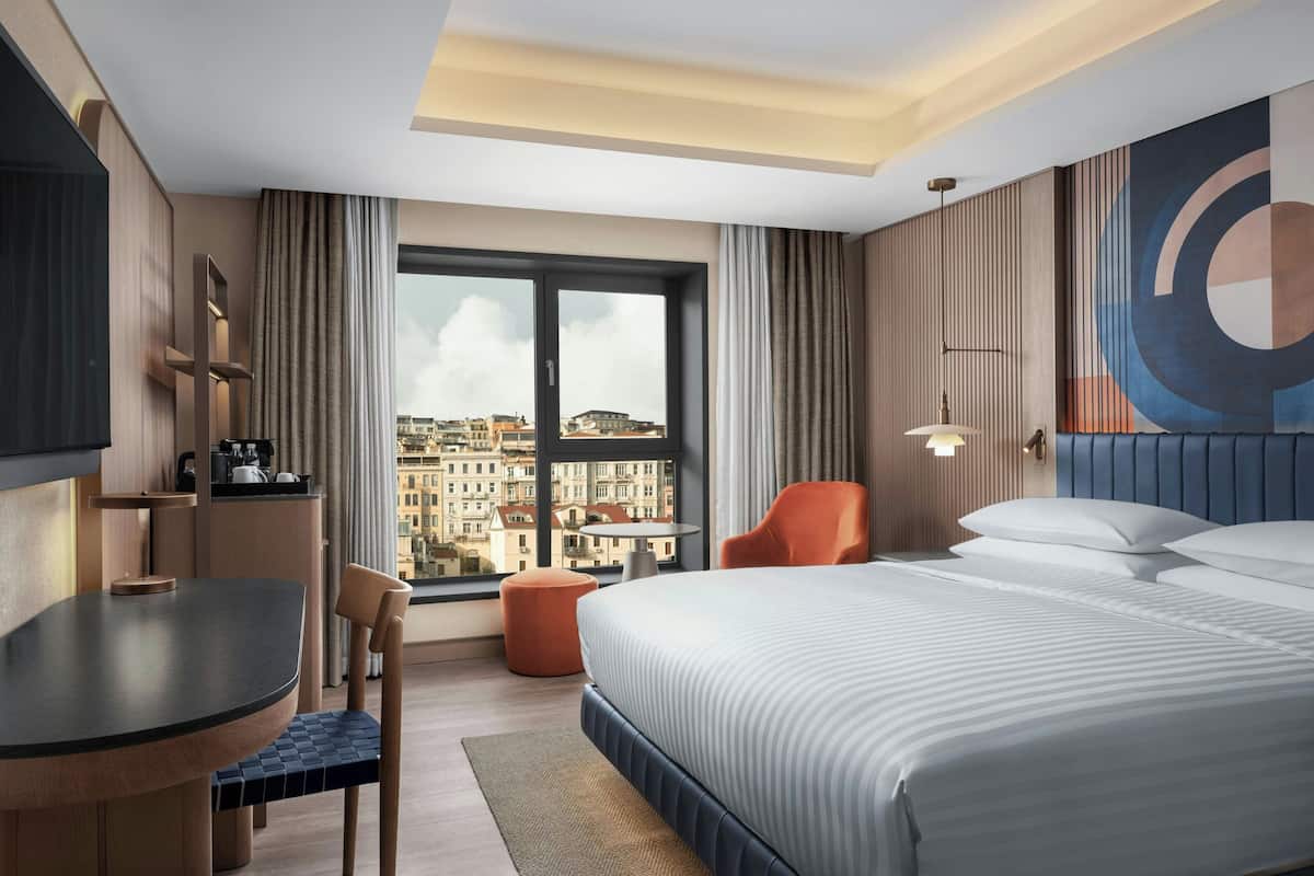 A modern hotel room with a large bed, a dining table, a chair, a small ottoman, and a view of the city through a large window.