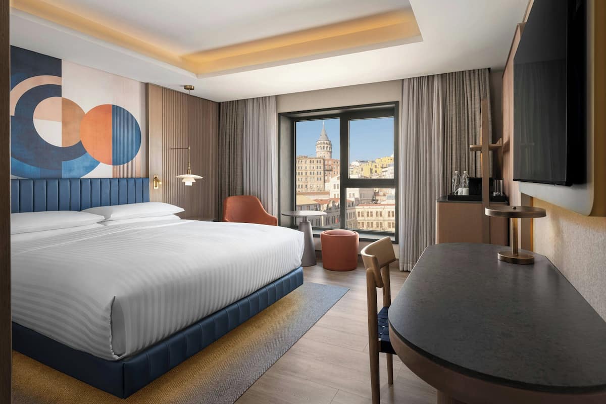 A hotel room with a large bed, a desk, a chair, a TV, and a view of a cityscape.