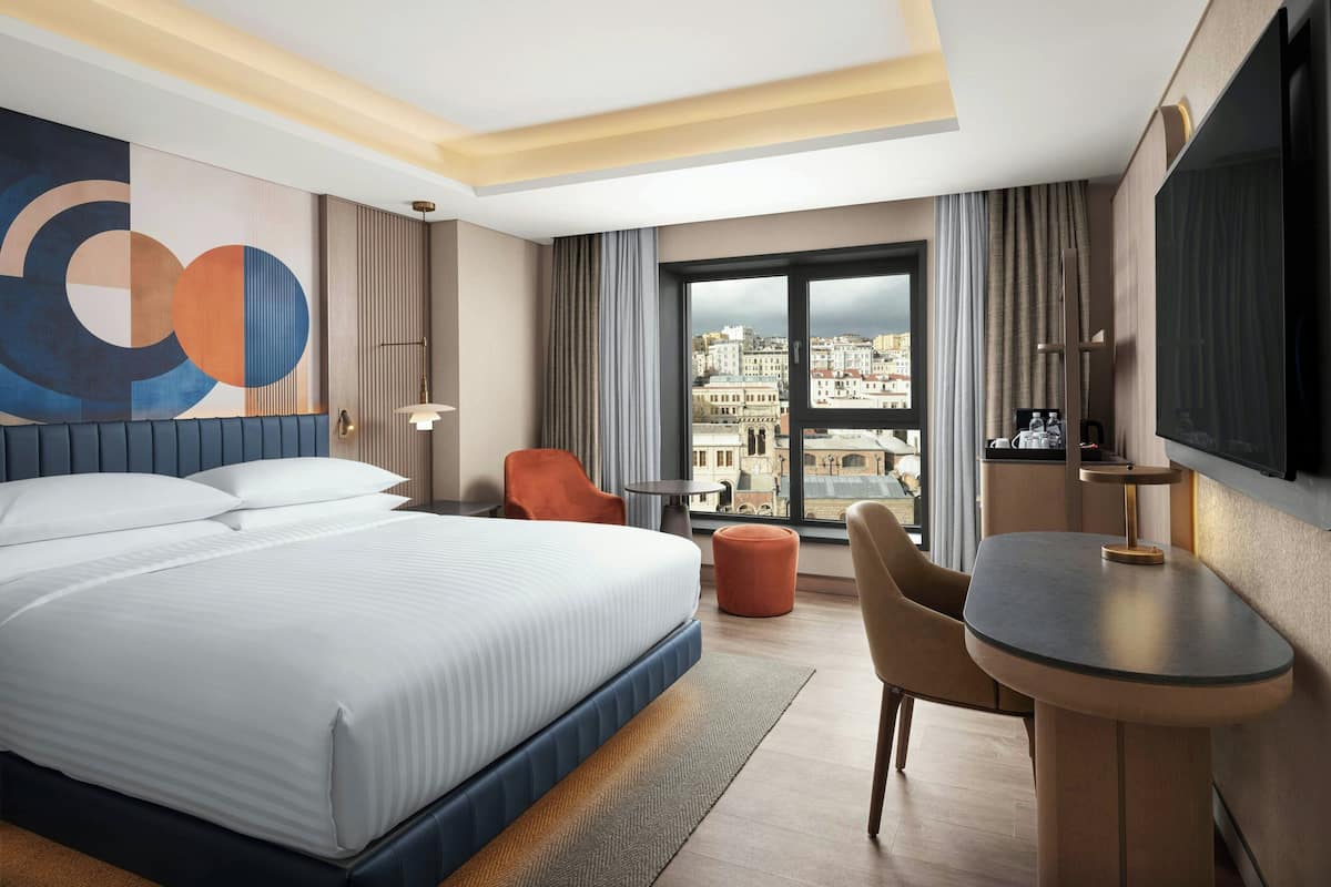 A hotel room with a large bed, a desk, and a TV. There is a view of buildings through the window.