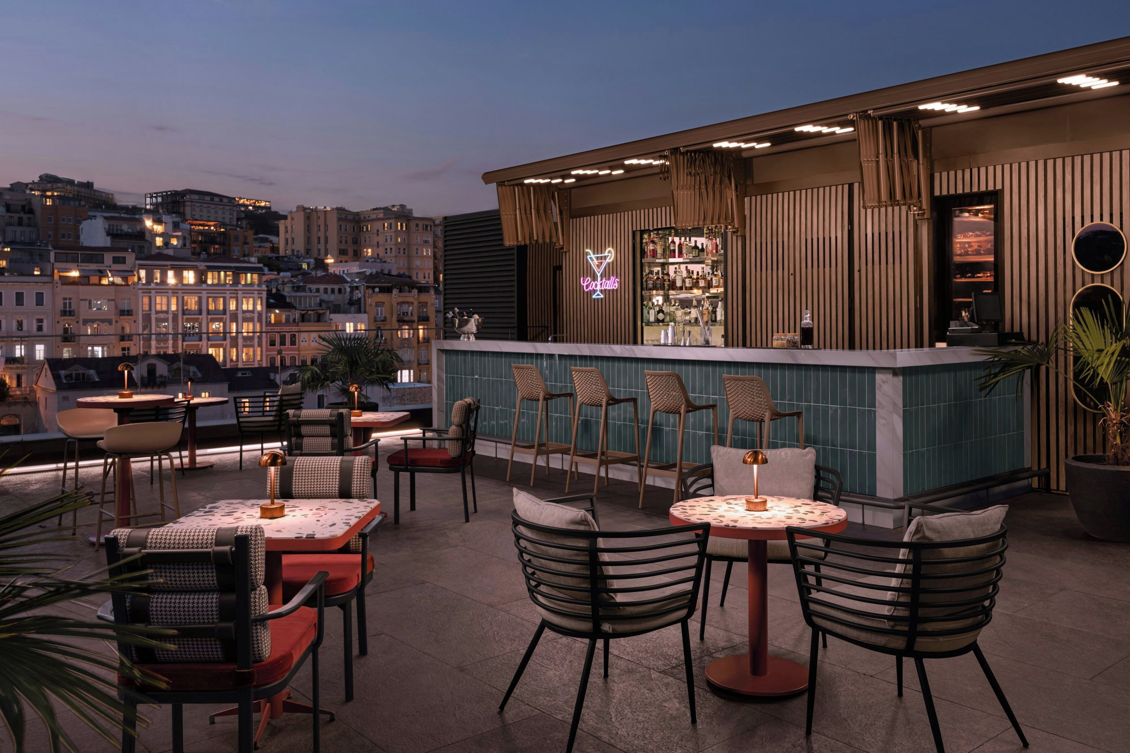 A rooftop bar with multiple tables and chairs, a bar counter with a neon sign, and a cityscape view in the background.