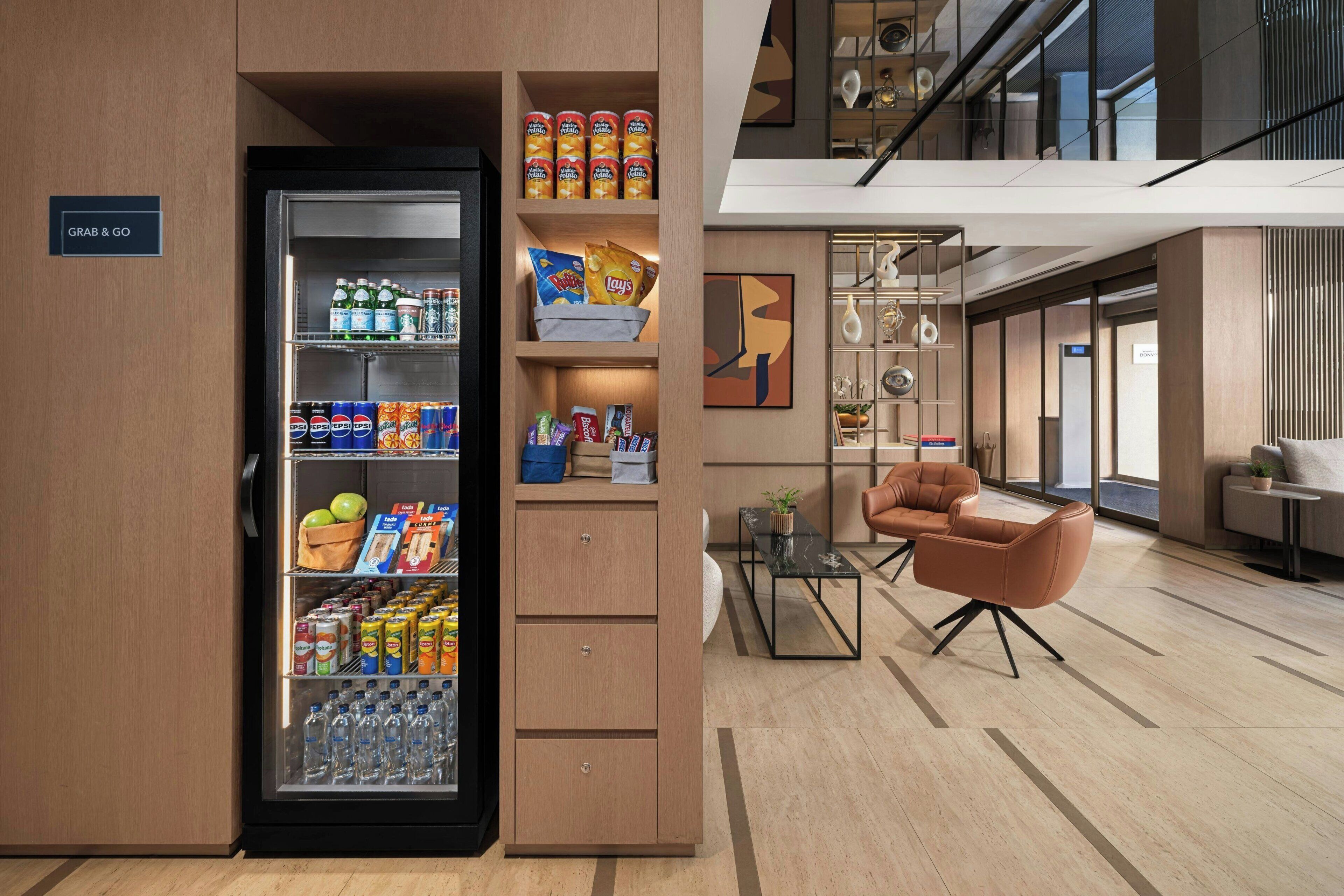 A modern hotel lobby with a 'Grab & Go' refrigerator stocked with snacks and drinks, a seating area with a sofa and armchairs, and a view of the upper floor.