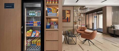 A modern hotel lobby with a 'Grab & Go' refrigerator stocked with snacks and drinks, a seating area with a sofa and armchairs, and a view of the upper floor.