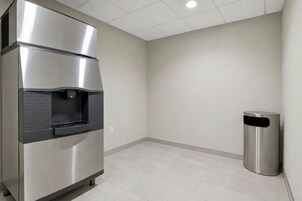 Un dispensador de bebidas de acero inoxidable y un cubo de basura en una habitación con suelo de baldosas y una pared lisa.