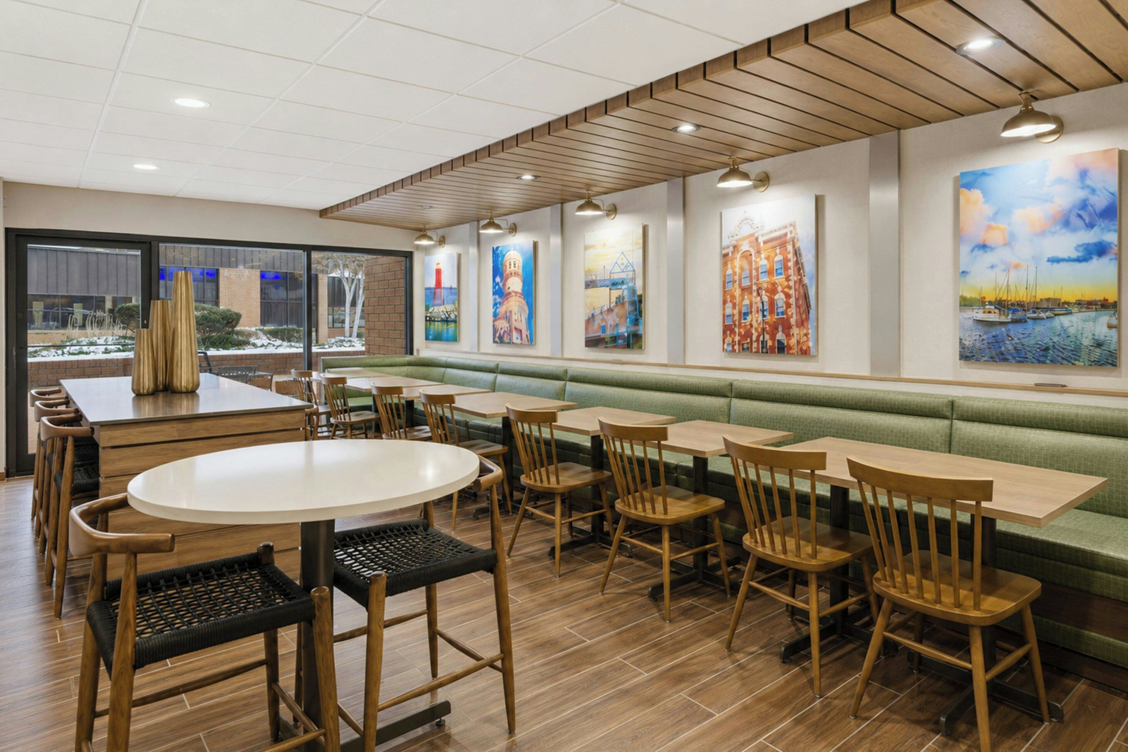 A restaurant interior with wooden tables and chairs, a green cushioned bench, and artwork on the walls.