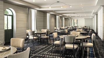 A spacious hotel dining area with round tables and chairs, large windows, and a patterned carpet.