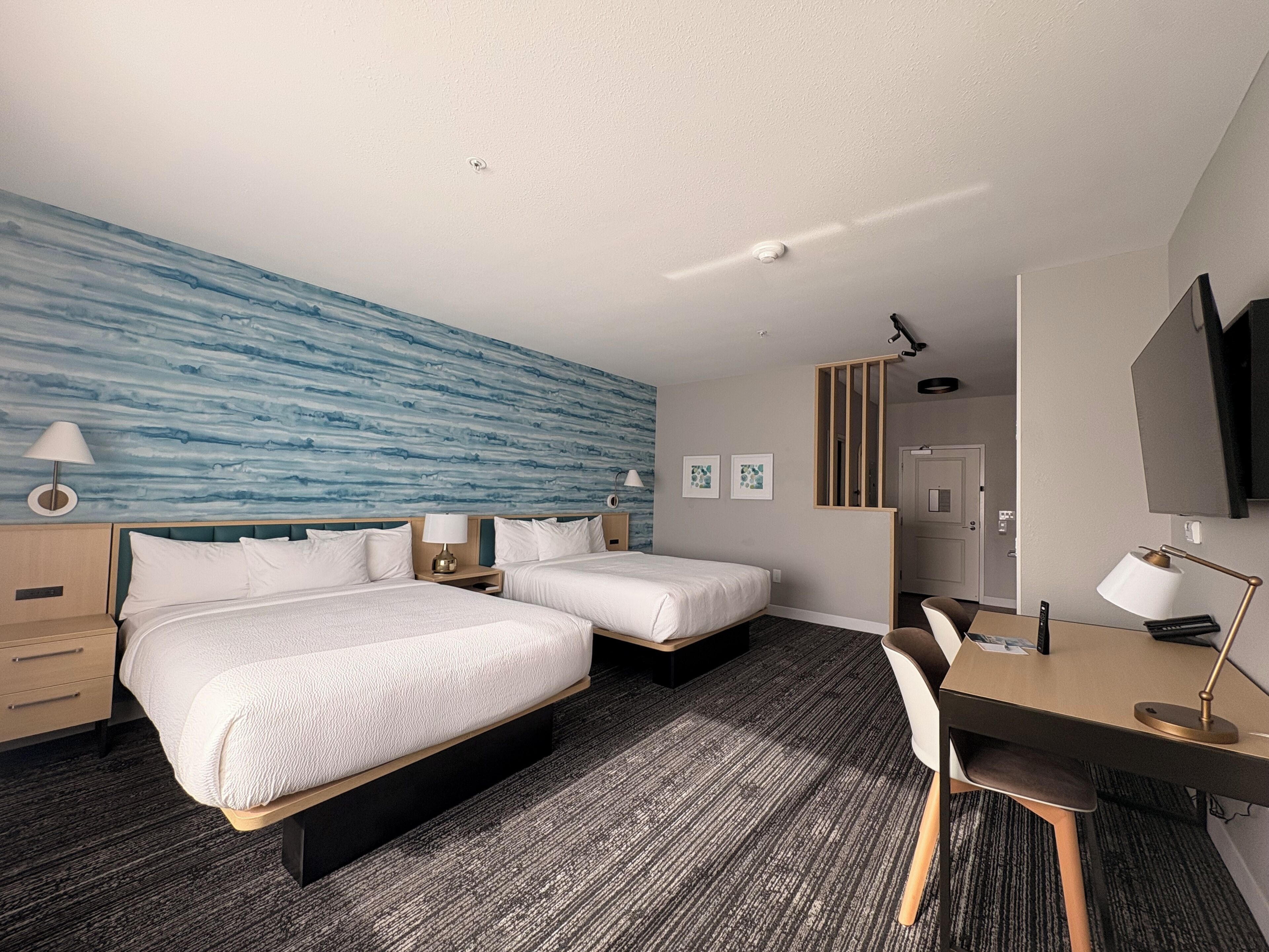 Habitación de hotel con dos camas, un escritorio, una silla, un televisor y una pared azul grande.