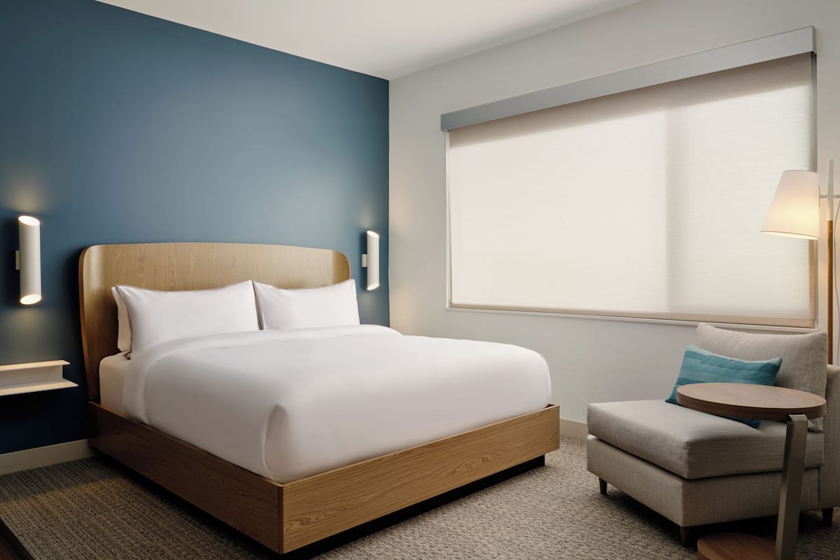 A modern hotel room with a wooden bed, a small armchair, a round side table, and a large window with blinds.