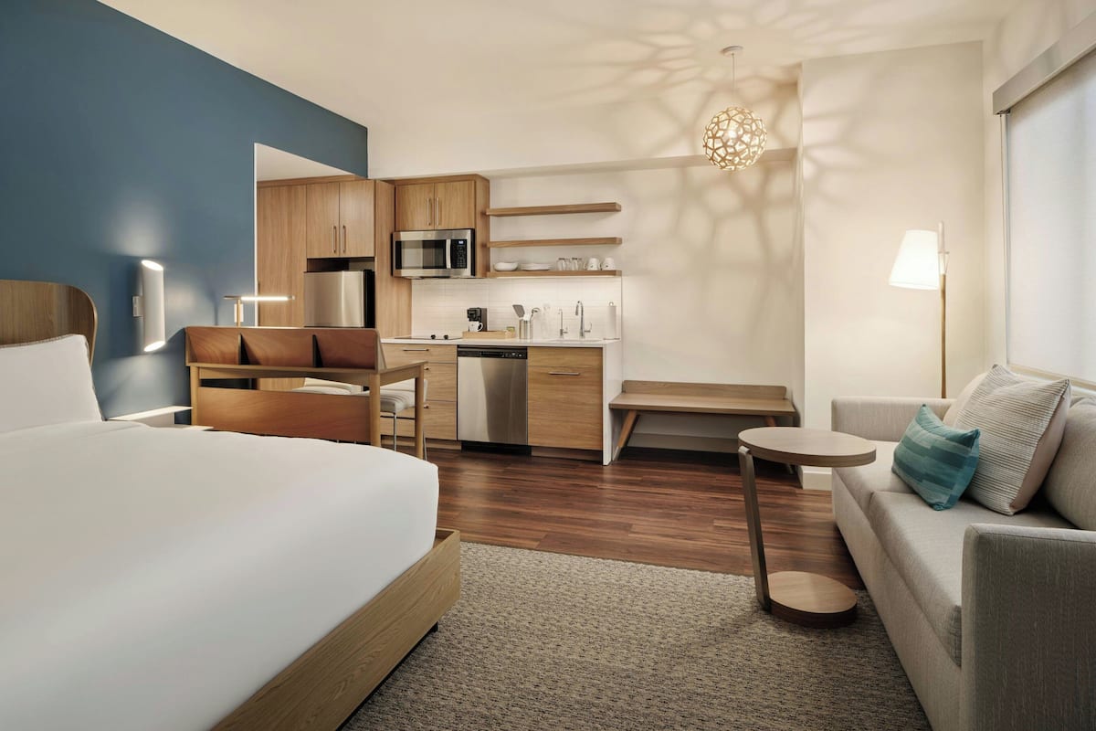 A modern hotel room with a large bed, a sofa, a small kitchen area, and a dining table.