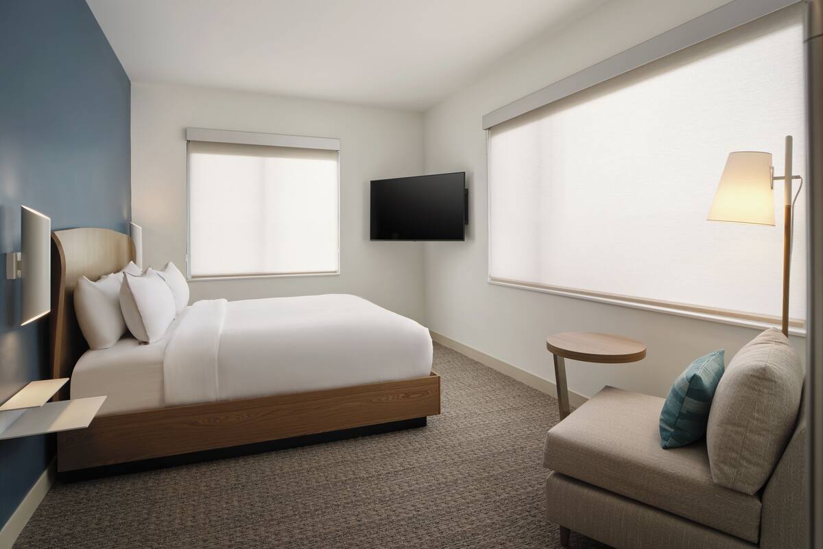 A modern hotel room with a bed, a TV mounted on the wall, a small round table, and a sofa with pillows.