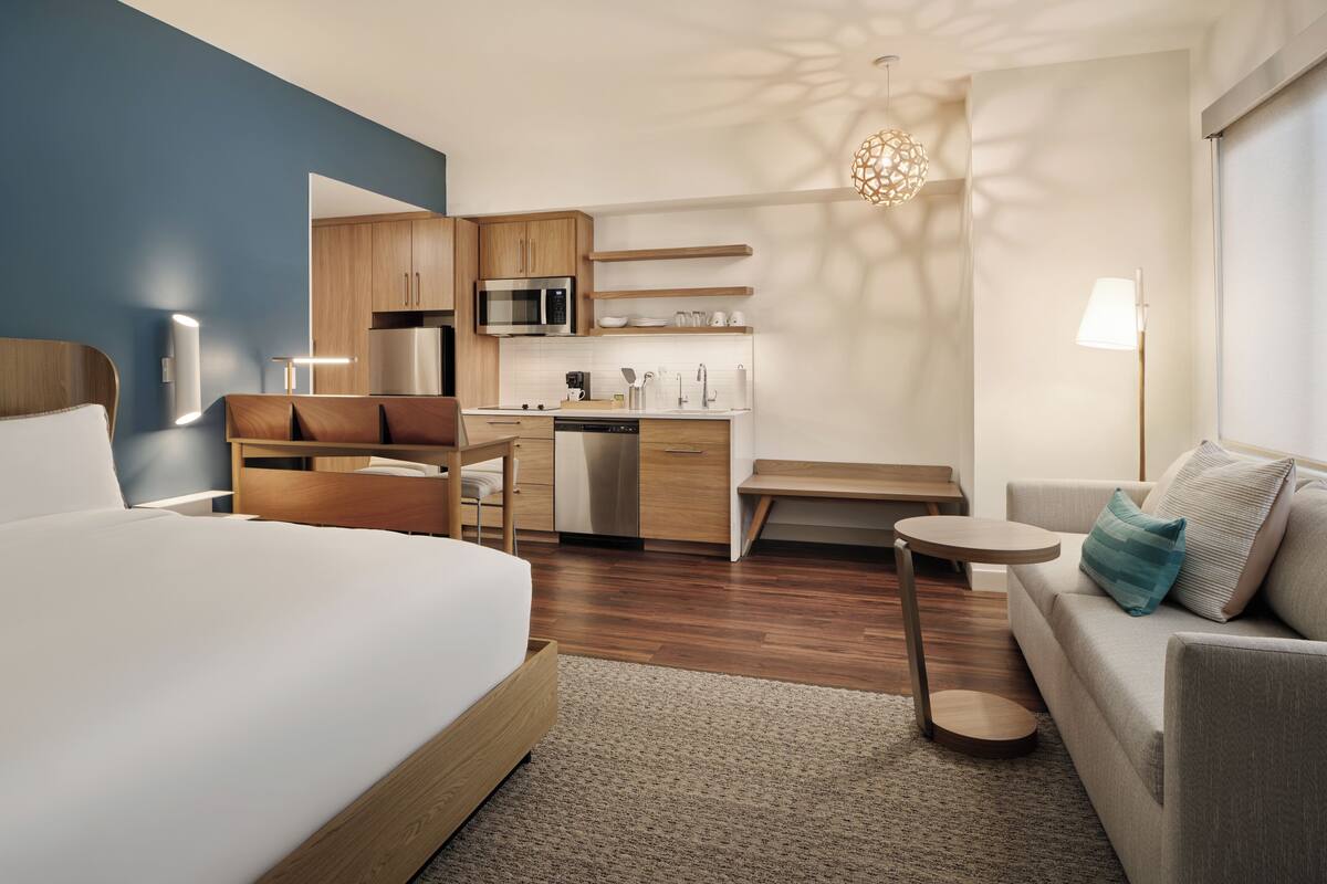 A modern hotel room with a large bed, a sofa, a small dining table, a kitchen area, and a bathroom.
