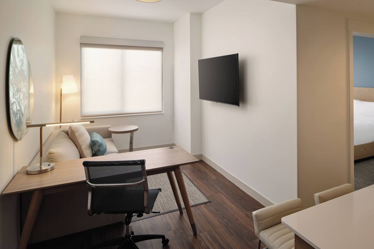 A modern hotel room with a wooden desk, a black office chair, a flat-screen TV mounted on the wall, a window with blinds, a sofa with cushions, and a small round table.