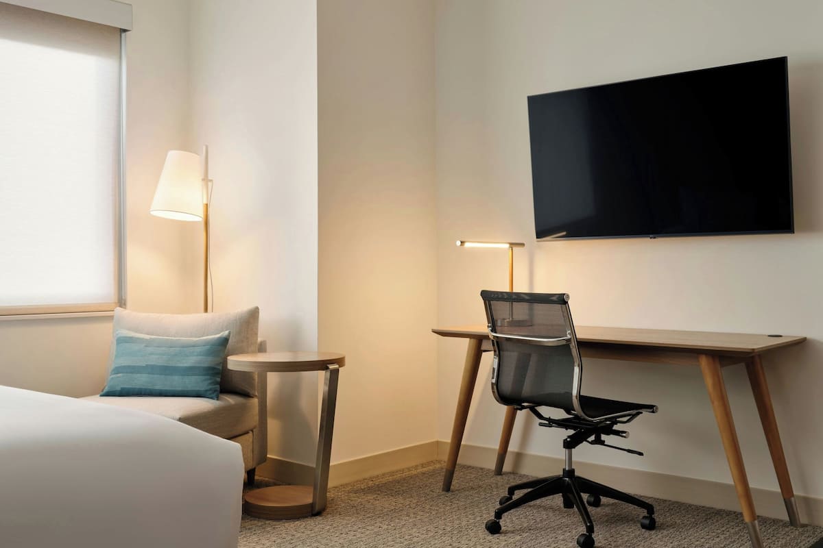 A hotel room with a bed, a desk, a chair, a TV, and a lamp.
