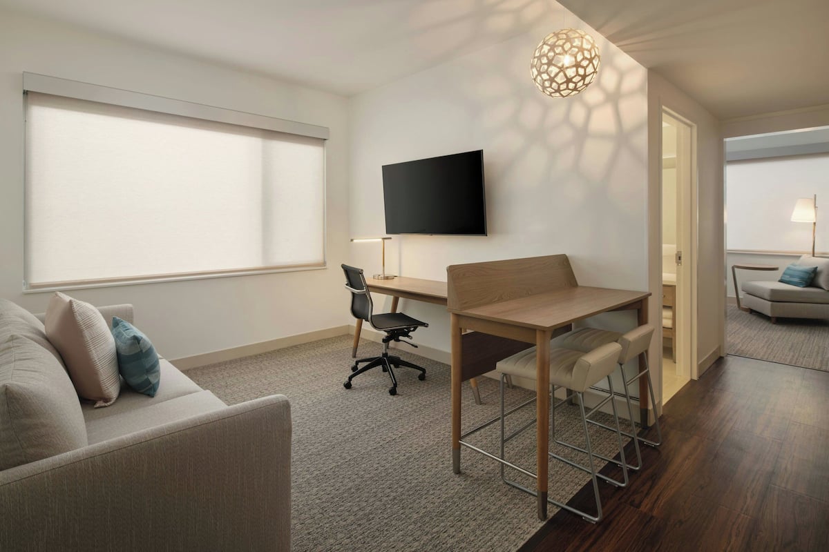 A modern hotel room with a flat-screen TV, a desk with a chair, a sofa, and a dining area with chairs.