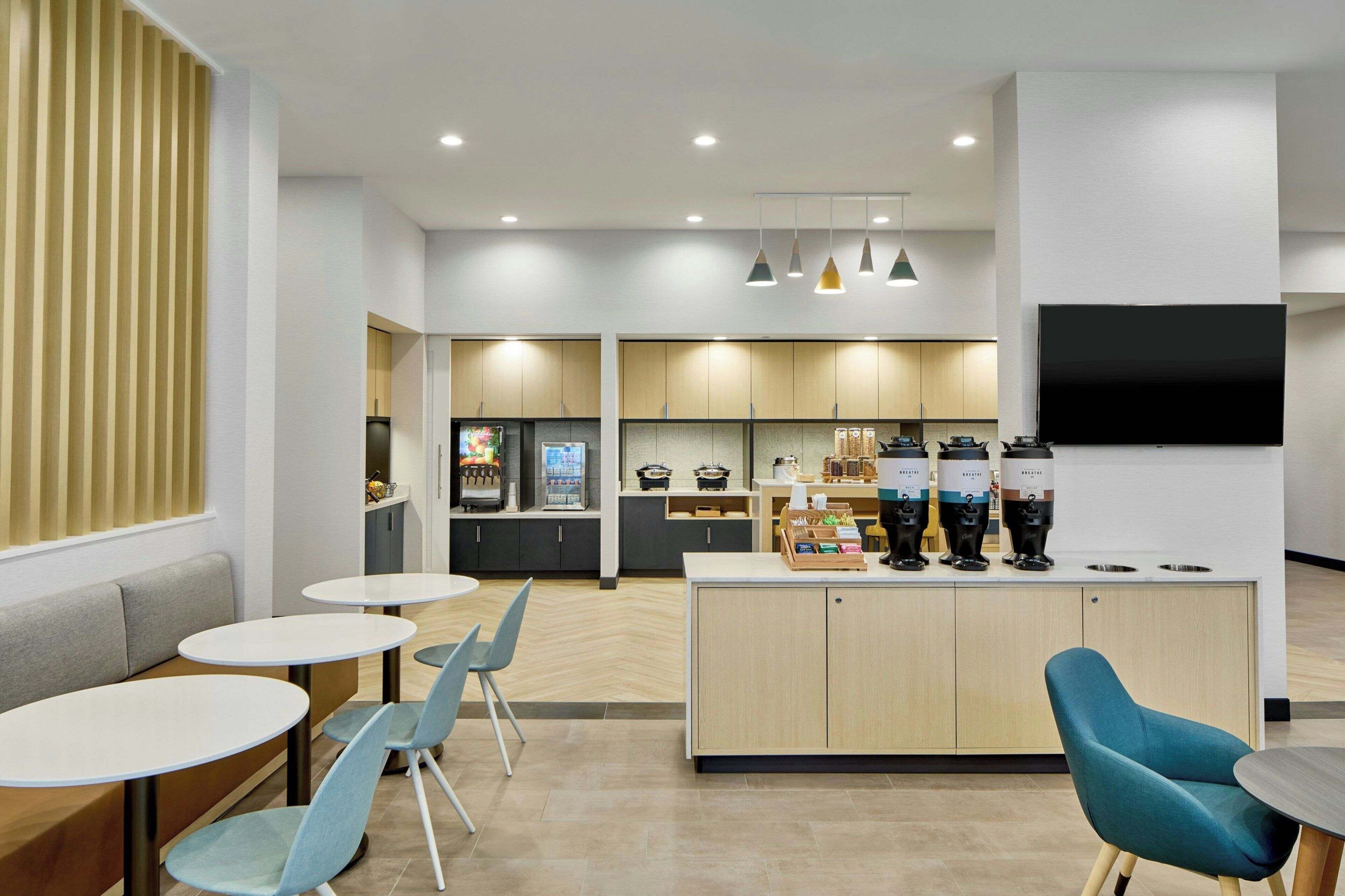 Un interior moderno con mesas redondas, sillas azules, una zona de cocina y un televisor de pantalla plana.