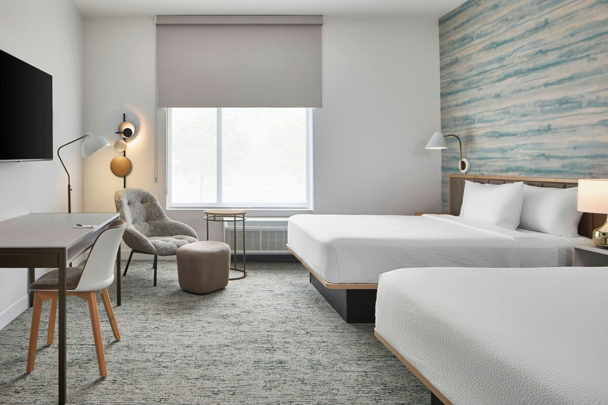 Une chambre d’hôtel avec deux lits, un bureau, une chaise, une petite table et une grande fenêtre.