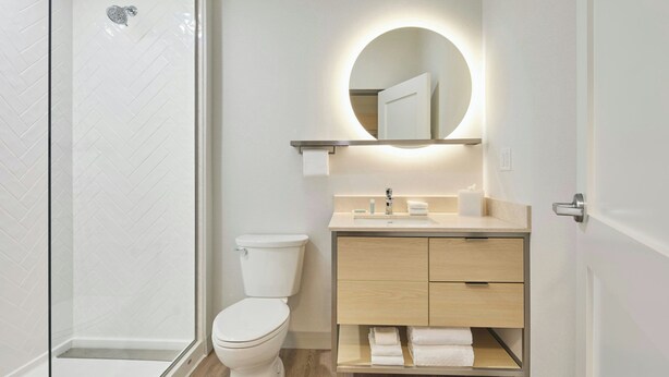 A modern bathroom with a glass-enclosed shower, a toilet, a wooden vanity with a mirror, and a shelf with towels. - Towneplace Suites by Marriott Tullahoma (Tullahoma)