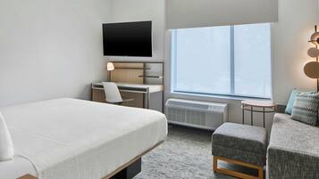 Habitación de hotel con cama, escritorio, silla, televisor, ventana y una mesita con lámpara.