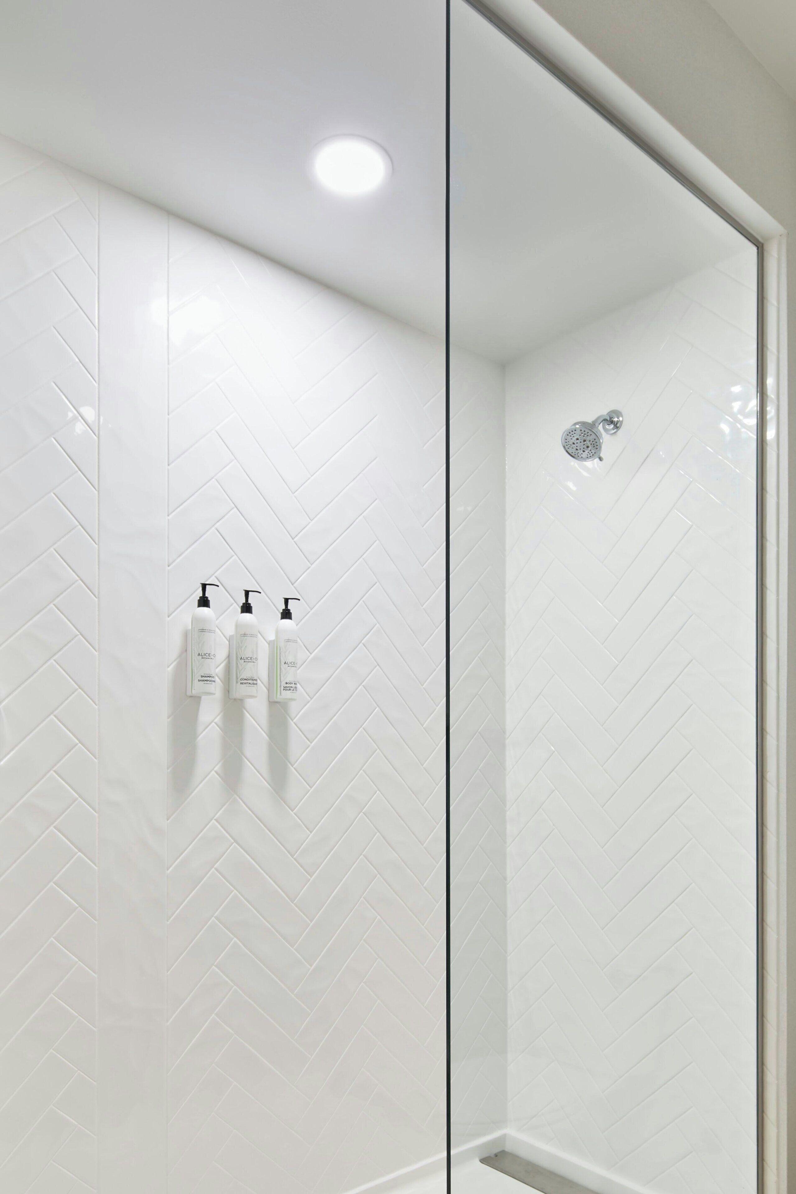 A shower with a glass door, white herringbone tiles, and three bottles of shampoo on the wall.