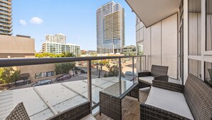 Un balcón con mobiliario de mimbre y barandilla de vidrio que da a una vista panorámica de la ciudad con edificios modernos.