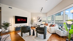 Una sala de estar moderna con un televisor de pantalla plana mostrando 'NETFLIX', un sofá, dos sillones, una mesa de centro y un ventanal con vistas a la ciudad.