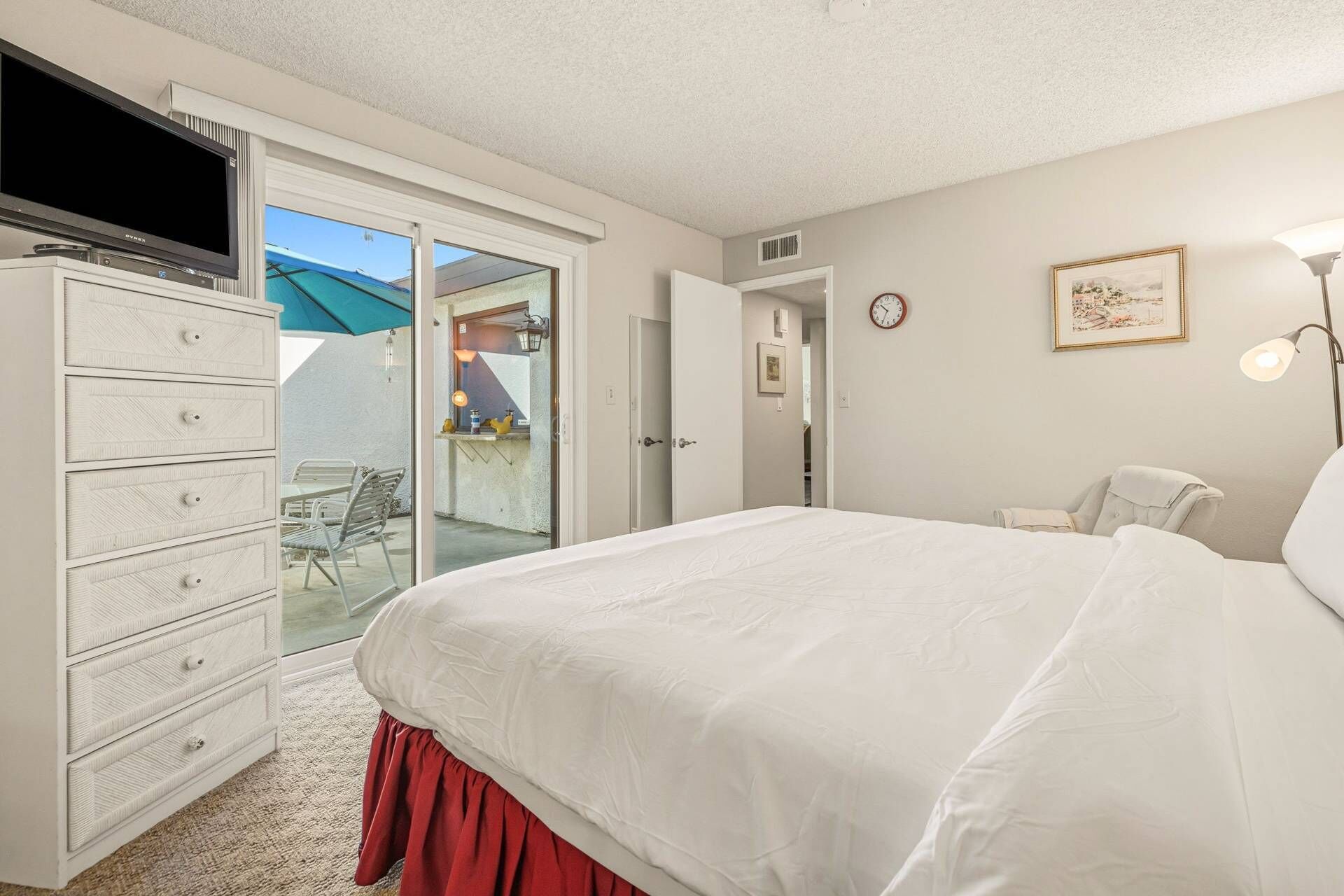 A bedroom with a large bed, a dresser, a TV, and a door leading to a balcony.