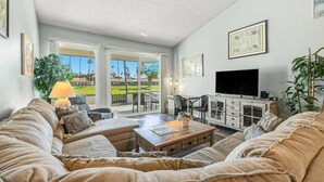 A spacious living room with a large sofa set, a coffee table, and a TV on a wooden cabinet. There is a balcony with a view of palm trees and a golf course. - Gorgeous Two Bedroom in Rancho Las Palmas (Rancho Mirage)