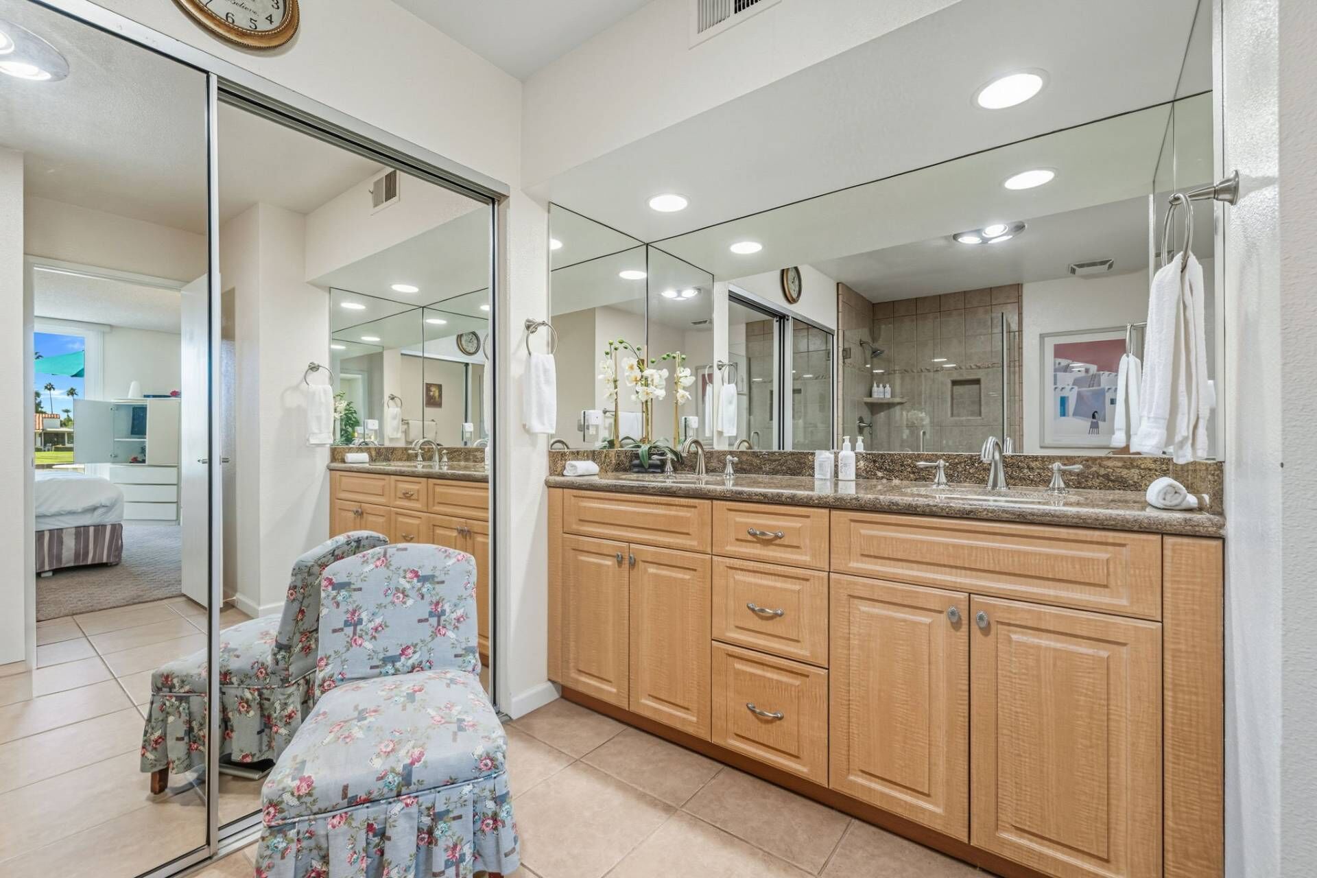 A bathroom with a large mirror, wooden cabinets, a floral chair, and a visible shower area.