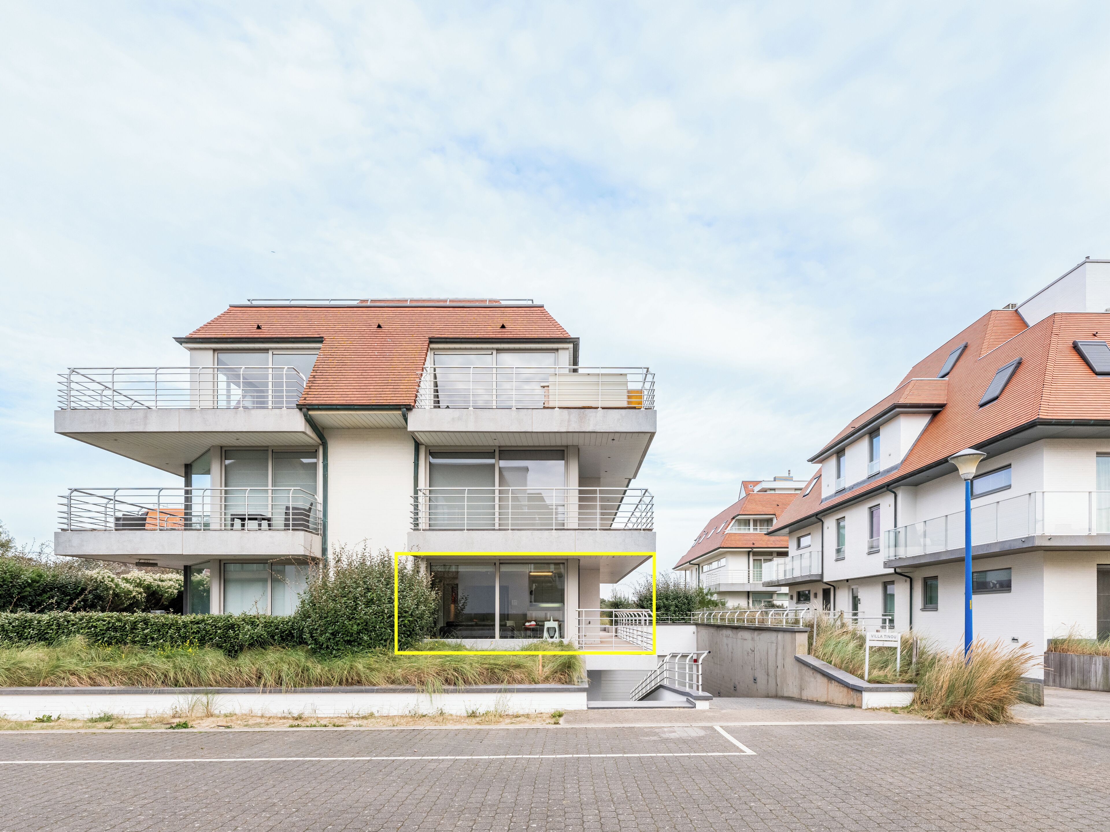 A modern multi-story residential building with balconies, a paved driveway, and a clear sky.