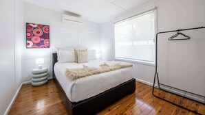 Une chambre à coucher comprenant un lit, un climatiseur fixé au mur, une fenêtre avec des stores et un porte-manteau fixé au mur.