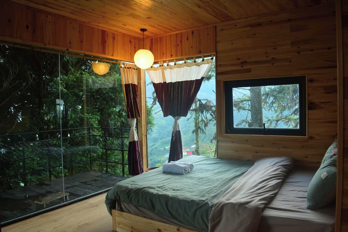 A wooden cabin bedroom with a large window, a bed, and a hanging light fixture.
