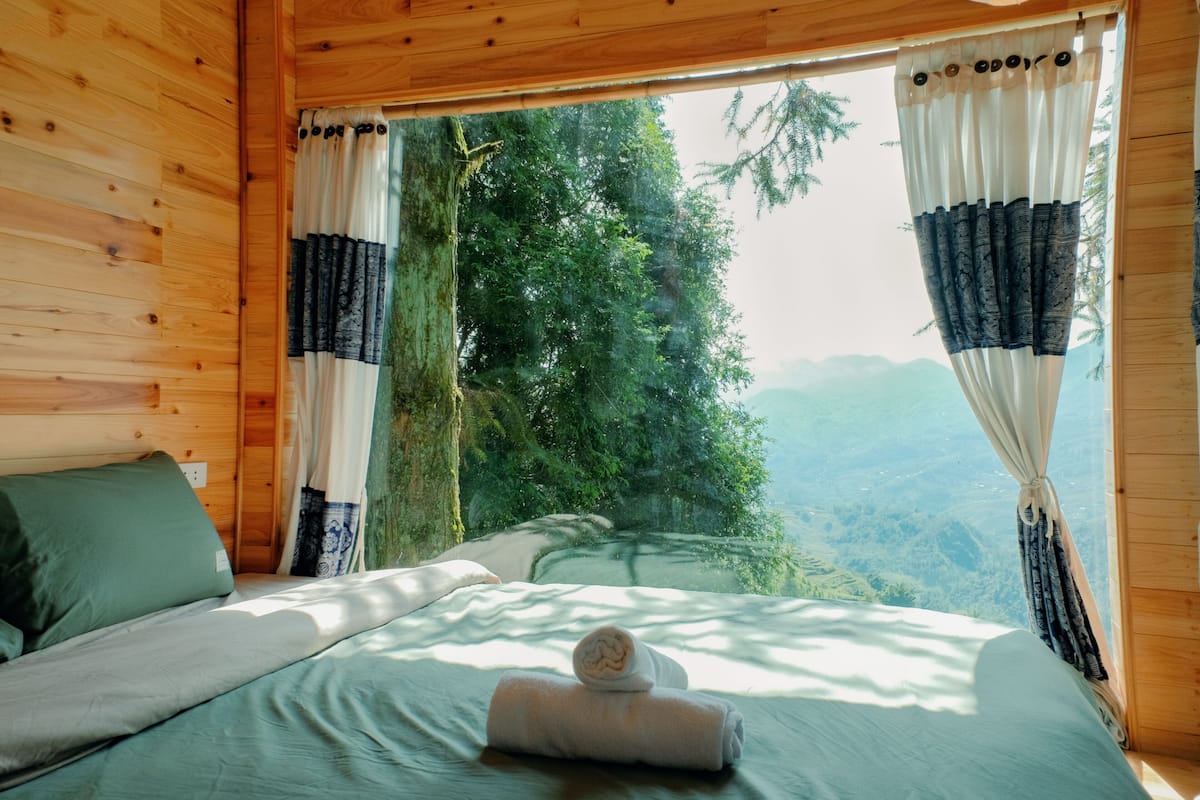 A wooden cabin bedroom with a bed, curtains, and a view of a forested landscape.