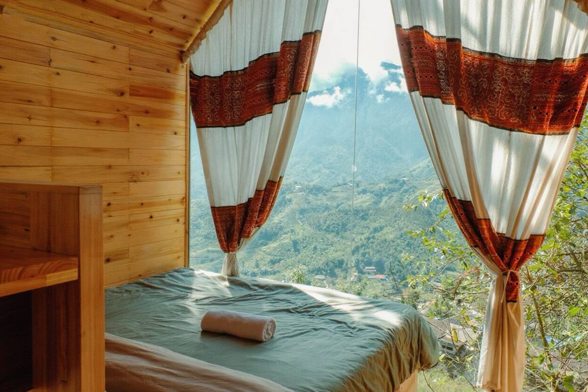 A wooden cabin room with a bed, curtains, and a view of a mountainous landscape.