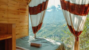 A wooden cabin room with a bed, curtains, and a view of a mountainous landscape.
