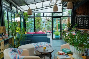 Amplio espacio interior con un gran ventanal que da a un jardín exterior, con un sofá azul, una pequeña mesa redonda y diversas plantas.