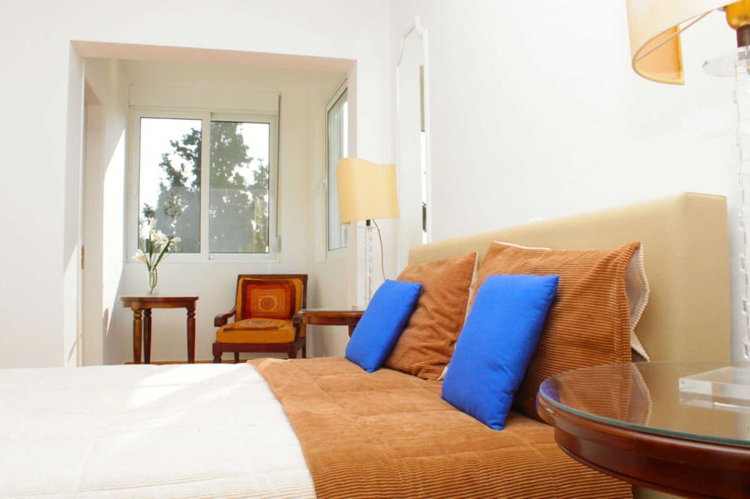 Une chambre d’hôtel avec un lit, une table ronde en verre, une lampe, une chaise et une fenêtre donnant sur des arbres.
