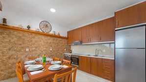 A kitchen with wooden cabinets, a stainless steel refrigerator, a stone wall, and a dining table set for four. - The Rhodes South Escape Beach (Rhodes)