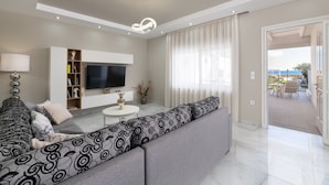 A modern living room with a large sofa, a flat-screen TV, and a dining area with outdoor seating. - The Cottage South Escape With Beach Front Pool (Rhodes)