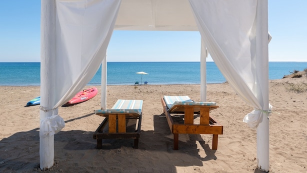 A beach setting with a white canopy structure, two wooden lounge chairs, and a red kayak on the sand. - The Cottage South Escape With Beach Front Pool (Rhodes)