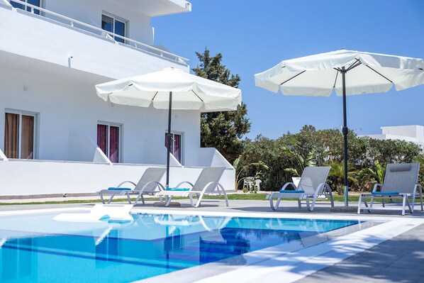 A pool area with white lounge chairs and umbrellas, a clear blue pool, and a white building with windows and a balcony. - Room 12 Bellezza Studios Groundfloor 12 (Rhodes)