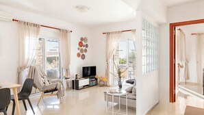 Un salon moderne avec un canapé, un téléviseur et une table basse, un coin repas avec des chaises, et de grandes fenêtres avec des rideaux.