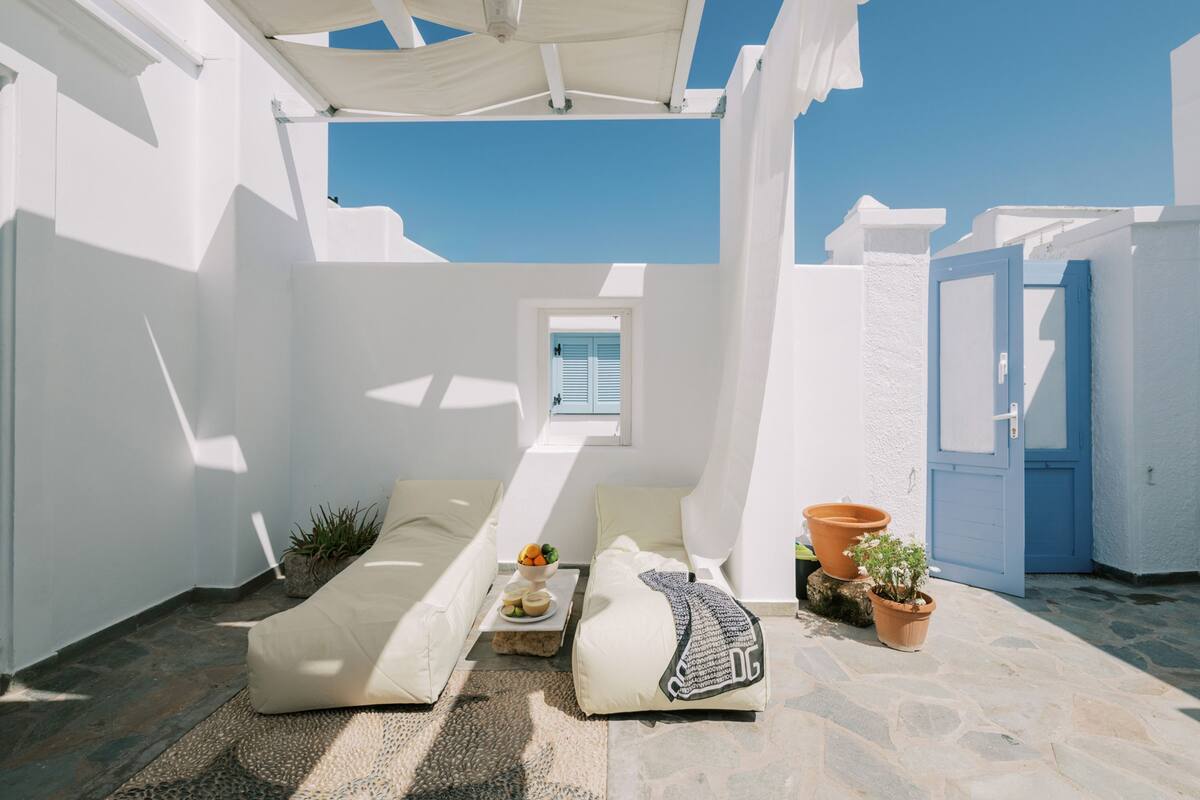 Un área de estar al aire libre iluminada por el sol, con una pared blanca, una puerta azul y un ventanal con contraventanas.