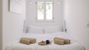 Cama bem arrumada com lençóis brancos, janela com vista e um quadro na parede.