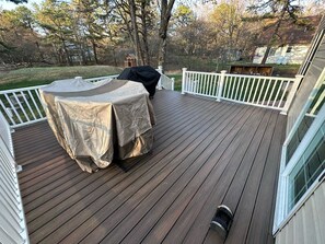 Una terraza de madera con una parrilla cubierta y una mesa, rodeada de barandas blancas.