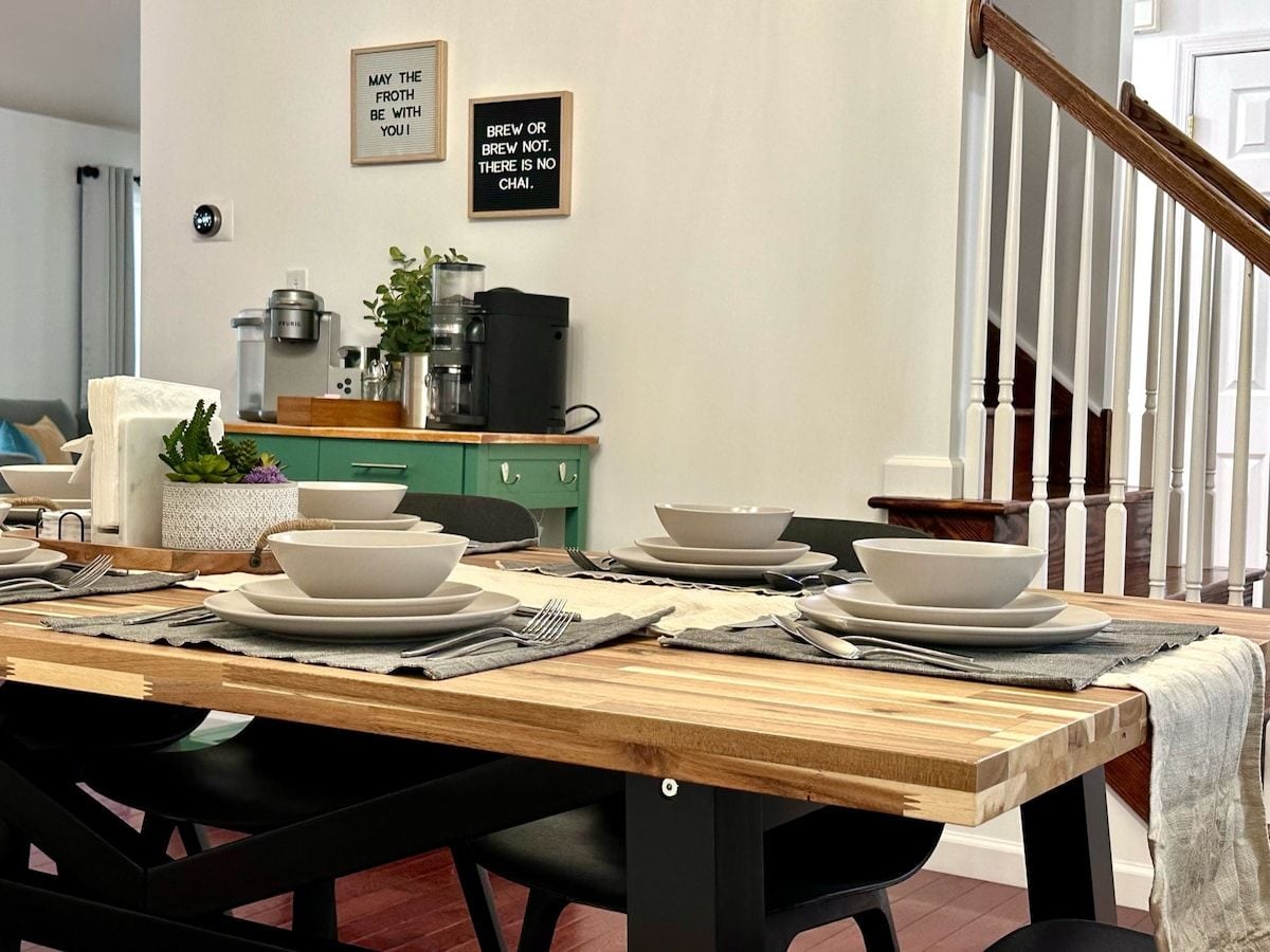 A dining table set with white plates, bowls, and cutlery, with a coffee machine and a plant in the background.