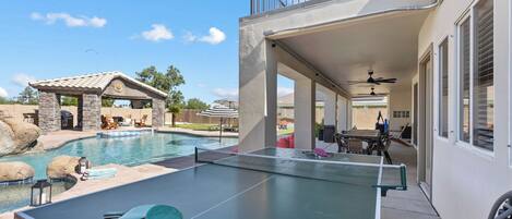 Une table de billard avec une raquette de tennis, un espace extérieur aménagé avec du mobilier de jardin, et une vue sur une piscine et un bâtiment doté d’un balcon.